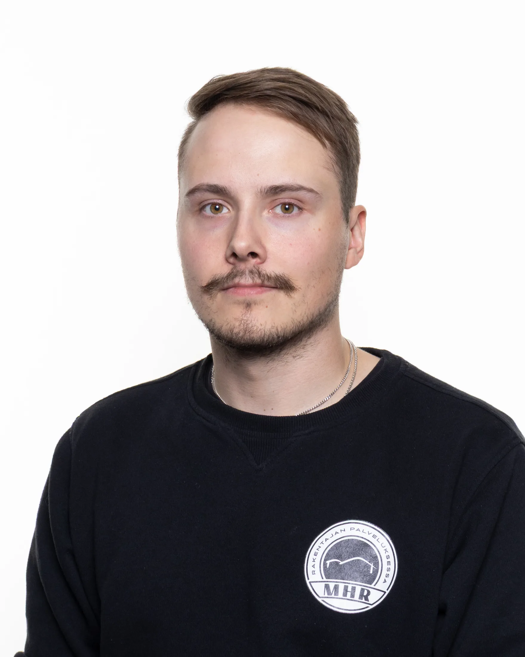 Muotokuva nuoresta miehestä, jolla on ruskeat hiukset ja viikset, mustassa paidassa, jossa on valkoinen logo, valkoista taustaa vasten.